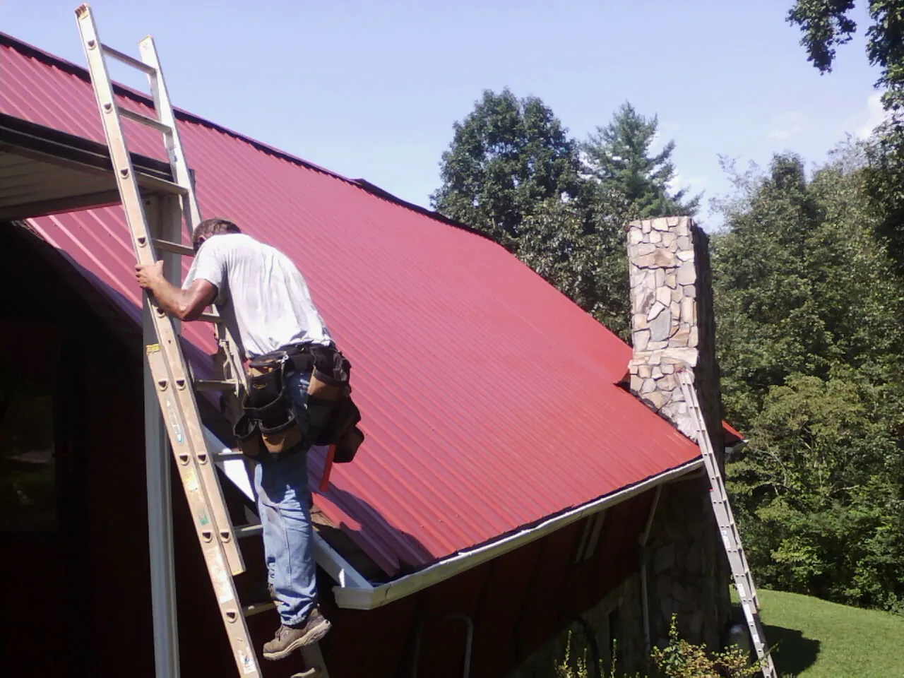 Roofer on a ladder beside a red metal roof and stone chimney — Soffit & Fascia Repair by Roofing Brothers in Waverly